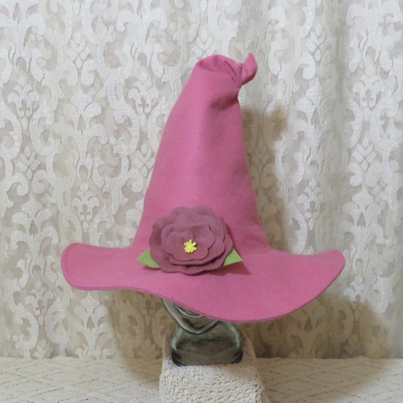 Puede incluir: Un sombrero de bruja de fieltro rosa con una gran flor de fieltro rosa en el borde. La flor tiene un centro amarillo.