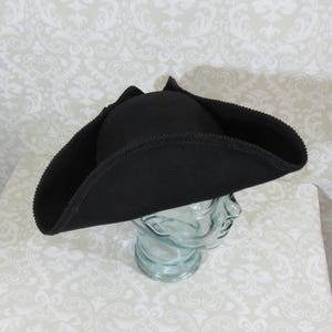 Puede incluir: Un sombrero tricornio negro con ala ancha y borde cosido decorativo. El sombrero se exhibe sobre una cabeza de cristal transparente. El dise&ntilde;o recuerda la vestimenta militar o pirata hist&oacute;rica.