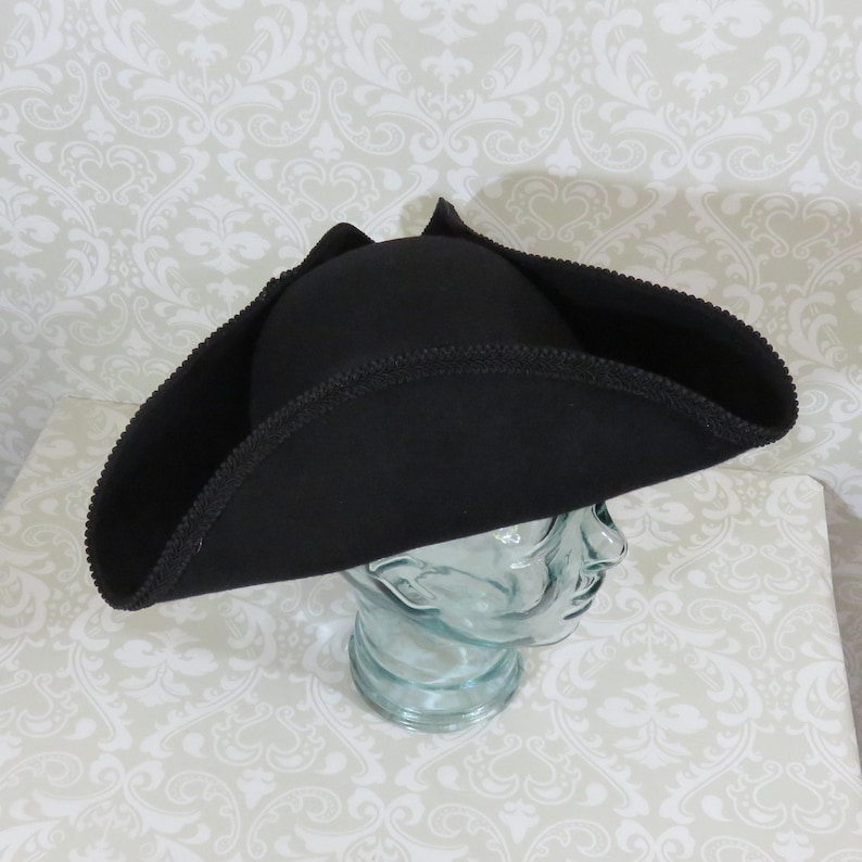 Puede incluir: Un sombrero tricornio negro con ala ancha y borde cosido decorativo. El sombrero se exhibe sobre una cabeza de cristal transparente. El dise&ntilde;o recuerda la vestimenta militar o pirata hist&oacute;rica.
