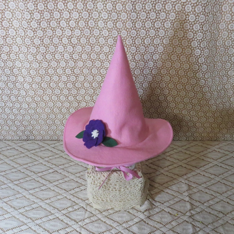 K&ouml;nnte beinhalten: Ein rosa Hexenhut mit spitzer Krone und breiter Krempe. Vorne ist eine lila Blume mit wei&szlig;em Zentrum und gr&uuml;nen Bl&auml;ttern befestigt. Der Hut wird auf einem mit Spitze bedeckten St&auml;nder pr&auml;sentiert.