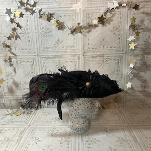 May include: A black hat with a peacock feather accent, a black flower, and a gold-colored button. The hat is displayed on a mannequin head. The background is a white wall with a star garland.