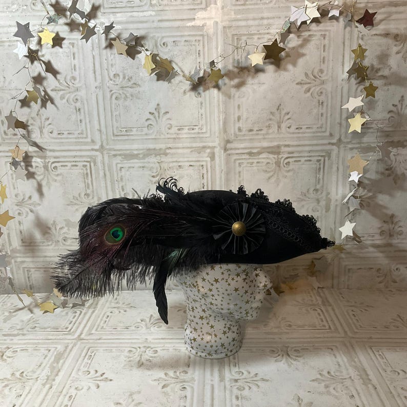 May include: A black hat with a peacock feather accent, a black flower, and a gold-colored button. The hat is displayed on a mannequin head. The background is a white wall with a star garland.