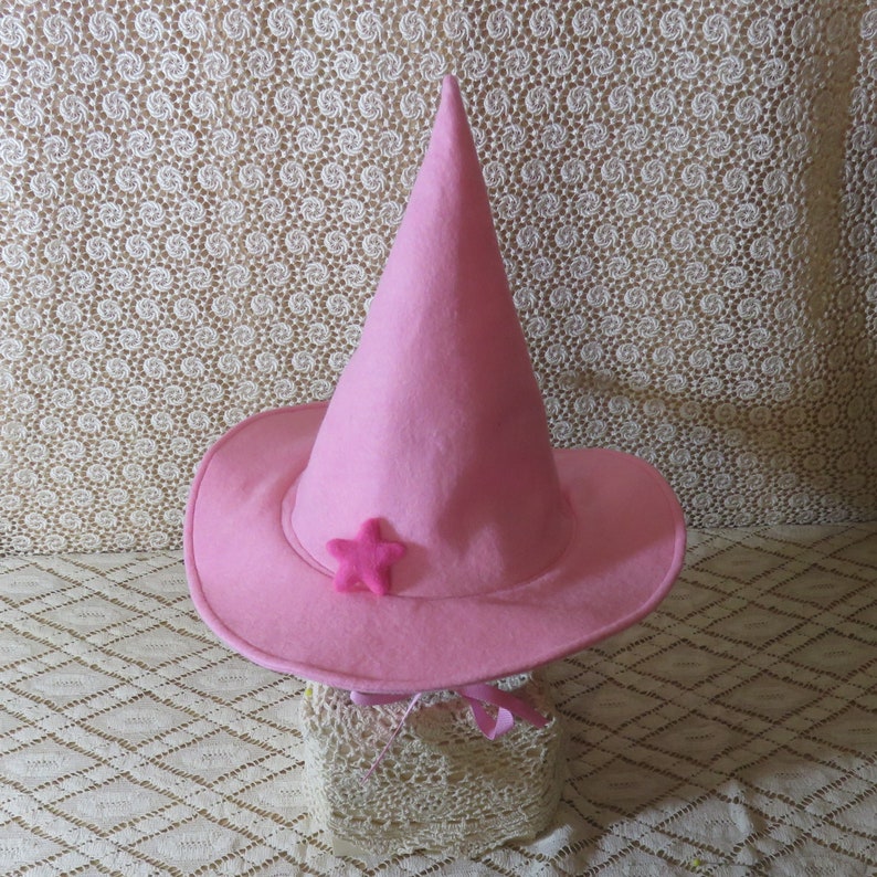 K&ouml;nnte beinhalten: Ein rosa Hexenhut mit breiter Krempe und spitzer Krone. Ein kleiner rosa Stern ist vorne befestigt. Der Hut ist aus Filz und hat ein Band um den Sockel gebunden.