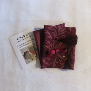 May include: A kit for making a Regency purse, with instructions, fabric, a tassel, ribbon, and a safety pin. The fabric is a burgundy color with a floral pattern.