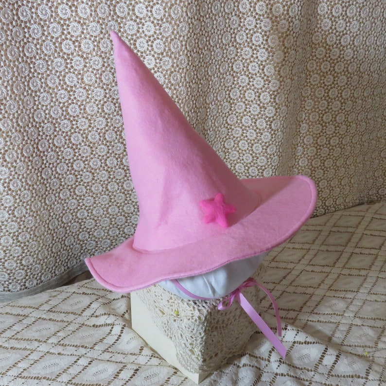 K&ouml;nnte beinhalten: Ein rosa Hexenhut mit spitzer Krone und breiter Krempe. Ein kleiner rosa Stern ist vorne befestigt. Rosa B&auml;nder sind an der Basis befestigt. Der Hut ist aus Filz.
