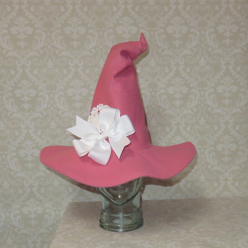 Puede incluir: Un sombrero de bruja rosa con un gran lazo de sat&eacute;n blanco y un acento de encaje blanco. El sombrero est&aacute; sentado en un soporte de exhibici&oacute;n de vidrio.