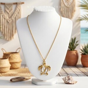 Puede incluir: Collar dorado con un colgante de tortuga, exhibido en un busto blanco. El colgante presenta una incrustación de nácar. El collar se presenta sobre un fondo de decoración de temática playera, incluyendo plantas y cestas tejidas.