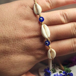 Könnte beinhalten: Ein goldfarbenes Armband mit drei weißen Muscheln und zwei blauen Evil-Eye-Perlen. Das Armband wird auf einer Hand gezeigt, mit einem verschwommenen Hintergrund aus Blumen.