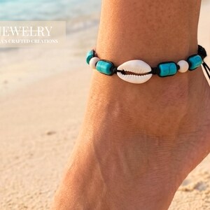 Könnte beinhalten: Fußkettchen mit einer weißen Muschelschale, türkisfarbenen und weißen Perlen an einer schwarzen Schnur. Das Fußkettchen wird an einem Fuß am Strand getragen. Der Text "JEWELRY" und "Mila's Crafted Creations" befindet sich in der oberen linken Ecke.