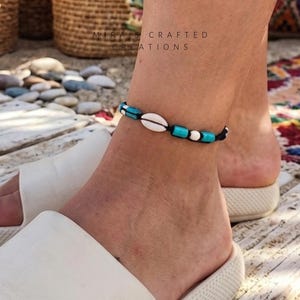 Könnte beinhalten: Fußkettchen mit einer weißen Muschel, türkisfarbenen Perlen und schwarzem Kordel. Das Fußkettchen wird an einem Knöchel mit weißen Sandalen getragen. Der Hintergrund umfasst Holzbretter, Steine und geflochtene Körbe.