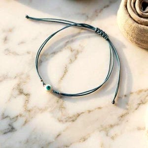 Pode incluir: Uma pulseira delicada com um cordão fino na cor verde-azulado e um pequeno pingente circular de olho grego com centro branco, azul e preto. A pulseira inclui um mecanismo de fecho com nó deslizante para ajuste de tamanho.
