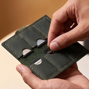 Könnte beinhalten: Ein Speicherkartenetui aus dunkelolivgrünem Leder mit mehreren vertikalen Fächern für SD-Karten, vernäht mit hellem Garn. Der geöffnete Organizer im Brieftaschenstil zeigt mehrere schwarze Speicherkarten, die in den einzelnen Taschen stecken.