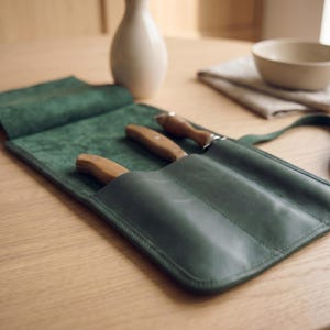 May include: A dark green leather knife roll featuring three individual slots, each containing a kitchen knife with a light brown wooden handle. The roll is partially open, revealing the smooth interior texture and the stitched pockets.