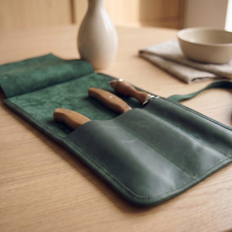 May include: A dark green leather knife roll featuring three individual slots, each containing a kitchen knife with a light brown wooden handle. The roll is partially open, revealing the smooth interior texture and the stitched pockets.