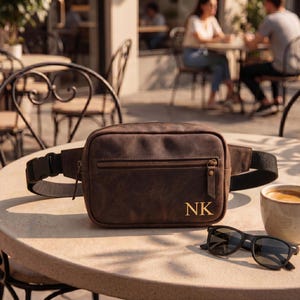 May include: A dark brown leather fanny pack featuring a horizontal zippered front pocket and the gold-colored initials NK embossed on the lower right corner. The bag includes a black adjustable nylon strap with a plastic side-release buckle.