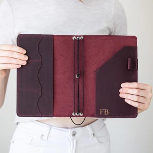 May include: A deep burgundy leather journal cover featuring three vertical card slots on the left interior panel, a diagonal pocket on the right, and three black elastic cords secured by metal eyelets. The initials FB are embossed on the right.