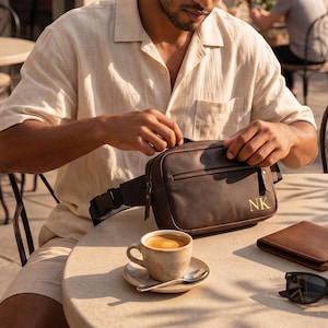 May include: A dark brown leather fanny pack featuring a front zippered pocket and the gold-embossed initials NK on the lower right corner. The bag includes a black adjustable strap with a plastic side-release buckle and matching leather zipper pulls.