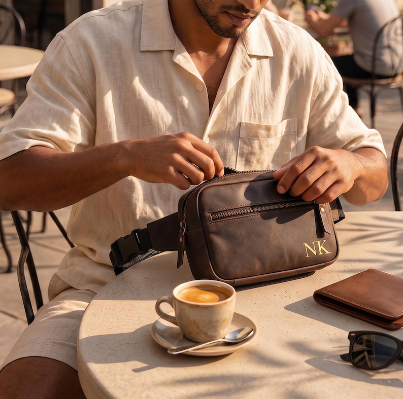 May include: A dark brown leather fanny pack featuring a front zippered pocket and the gold-embossed initials NK on the lower right corner. The bag includes a black adjustable strap with a plastic side-release buckle and matching leather zipper pulls.