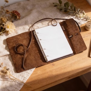 May include: Open brown leather journal with a spiral binding and blank white pages. The journal is open, revealing the interior pages, and is secured with a leather strap. A pen is tucked inside. The journal is on a wooden surface.