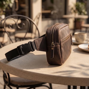 May include: A brown leather fanny pack featuring a rectangular main compartment with a top zipper, a smaller front zippered pocket, and a black nylon adjustable strap with a plastic side-release buckle. The bag has a smooth, matte finish.