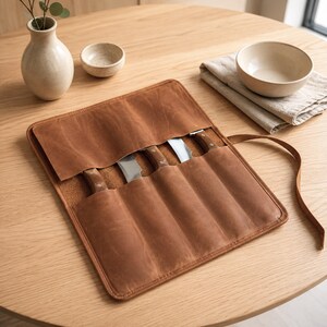 May include: A brown leather knife roll featuring five individual slots for storage. The roll is partially open, revealing the wooden handles and metallic blades of four knives tucked inside. A long leather strap is attached for securing the roll.