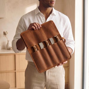 May include: A tan leather knife roll featuring multiple vertical slots for storing kitchen knives. The roll is constructed from smooth, matte-finish leather with visible stitching along the edges and includes a flap to cover the knife handles.
