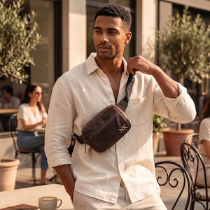 May include: A dark brown leather crossbody bag with a front zippered pocket and the gold-colored initials NK embossed on the lower right corner. The bag features a black adjustable strap and is worn across the chest over a light beige linen shirt.