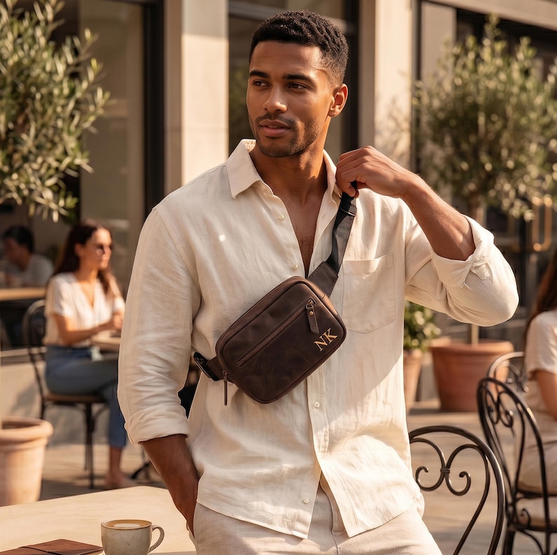 May include: A dark brown leather crossbody bag with a front zippered pocket and the gold-colored initials NK embossed on the lower right corner. The bag features a black adjustable strap and is worn across the chest over a light beige linen shirt.