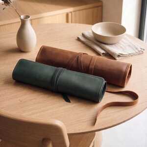 May include: Two cylindrical leather rolls, one in dark green and one in tan, each secured with a matching leather strap. The rolls feature visible stitching along the edges and a smooth, matte finish, presented as functional storage cases for items.