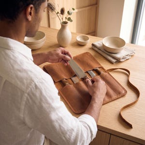 May include: A tan leather knife roll with multiple interior slots containing stainless steel kitchen knives with wooden handles. The roll features a long leather tie for securing the case when folded, showcasing a smooth, matte texture.