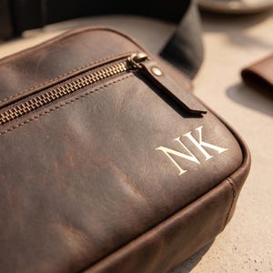 May include: A dark brown leather crossbody bag featuring a front zippered pocket with a metallic zipper pull. The bag is personalized with the embossed letters NK on the lower right corner and includes a black adjustable fabric strap.