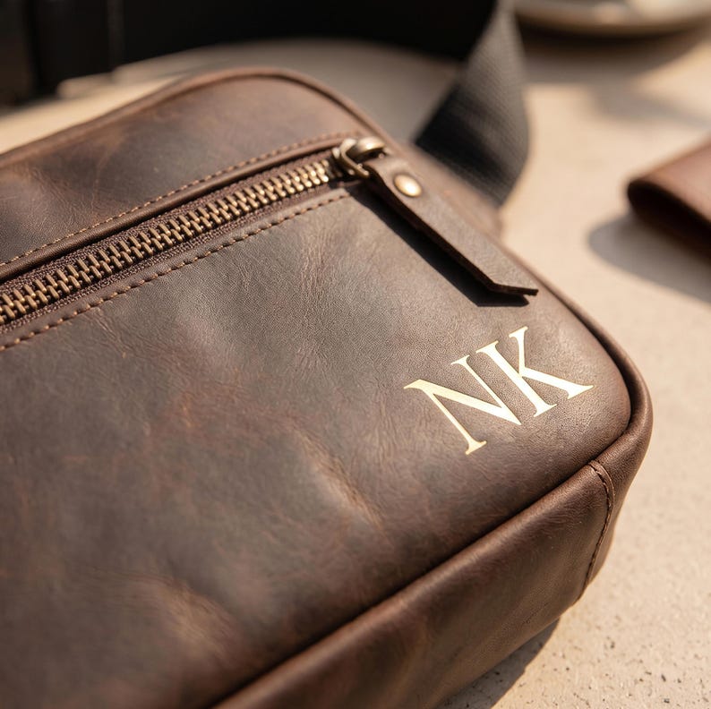 May include: A dark brown leather crossbody bag featuring a front zippered pocket with a metallic zipper pull. The bag is personalized with the embossed letters NK on the lower right corner and includes a black adjustable fabric strap.