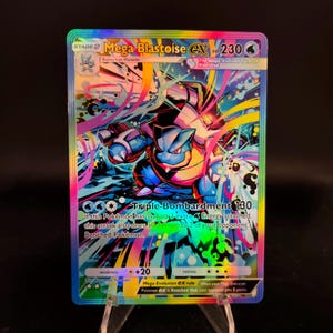 May include: A holographic Mega Blastoise EX Pokémon trading card. The card features a blue and black Blastoise character with colorful abstract art. Text includes "Mega Blastoise EX", "Triple Bombardment 130", and other game details.