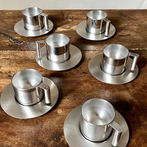 Op de afbeelding: Set van zes zilveren espresso kopjes en schoteltjes. De kopjes en schoteltjes zijn gemaakt van gepolijst metaal en hebben een eenvoudig, modern ontwerp.