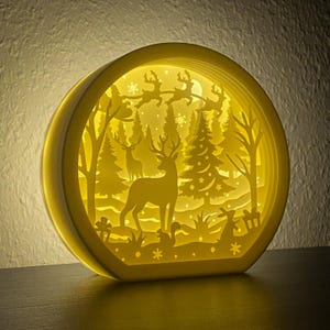 Könnte beinhalten: Eine weiße, runde, mehrschichtige, beleuchtete Papierschnitzerei, die eine Winterlandschaft mit einem Reh, einem Baum und fliegenden Rentieren zeigt. Die Szene wird von hinten beleuchtet und erzeugt ein warmes Leuchten.
