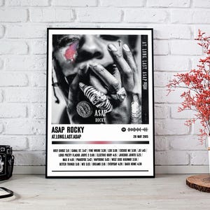 Puede incluir: Portada de álbum en blanco y negro para el álbum "At. Long. Last. ASAP" de ASAP Rocky. La portada presenta un primer plano de la cara de ASAP Rocky con un tatuaje de cruz en la frente. El título del álbum y la fecha de lanzamiento están impresos en blanco sobre un fondo negro.