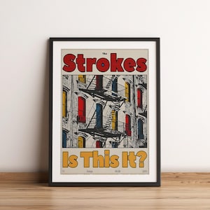 May include: Framed poster with the band name "The Strokes" in red, above the text "Is This It?" in yellow. The background depicts a building facade with colourful windows and fire escapes. The poster is in a black frame.