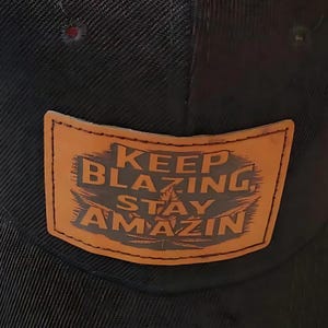 May include: Black baseball cap with a brown leather patch. The patch features the words "KEEP BLAZING STAY AMAZIN" in dark gray lettering. The cap has a curved brim and a mesh back.