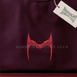 May include: A maroon crew neck sweatshirt with a red embroidered design and the text "MIDNIGHT DESIGN CLUB". A white tag with the same logo hangs from the collar. The sweatshirt is folded neatly on a red surface.
