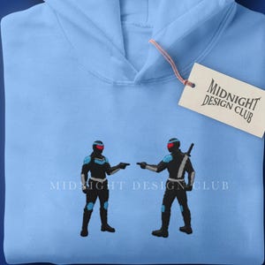 May include: Light blue hoodie with a graphic of two figures in black and blue armor, holding weapons. The hoodie has a hood and a tag that reads "Midnight Design Club". The text "Midnight Design Club" is also printed on the top left of the hoodie.