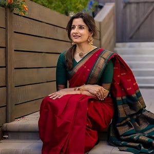 May include: A woman wearing a red sari with a green blouse, gold jewelry, and a gold necklace. The sari has a gold border and a green and gold border. The woman is sitting on stone steps.
