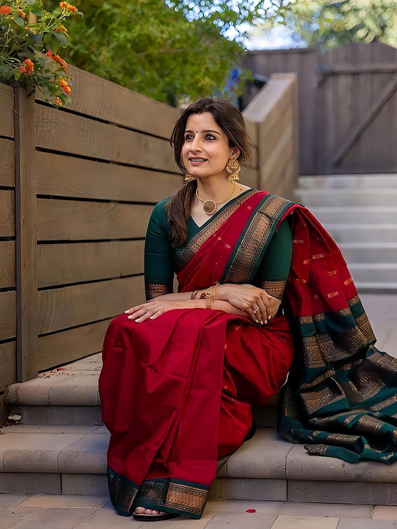May include: A woman wearing a red sari with a green blouse, gold jewelry, and a gold necklace. The sari has a gold border and a green and gold border. The woman is sitting on stone steps.