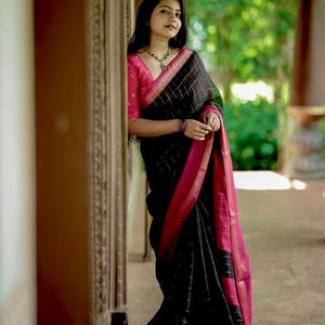 May include: A black saree featuring a subtle grid pattern, accented by a vibrant pink border and matching pink blouse. The garment is draped elegantly, showcasing the contrast between the dark fabric and the bright pink trim along the edges.
