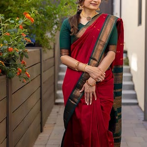 May include: A red saree with a green and gold border. The saree is draped over a green blouse. The image shows a woman wearing the saree, with gold jewelry, standing outside near a wooden fence and flowers.