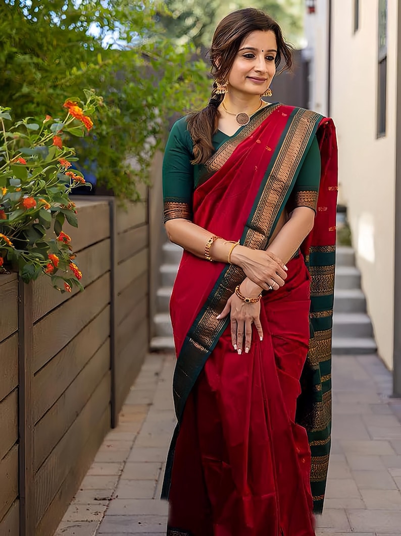 May include: A red saree with a green and gold border. The saree is draped over a green blouse. The image shows a woman wearing the saree, with gold jewelry, standing outside near a wooden fence and flowers.