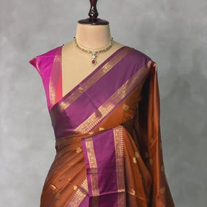 Puede incluir: Un sari tradicional en tonos cobre, morado y rosa, drapeado sobre un maniquí. El sari presenta detalles dorados a lo largo de los bordes y dispersos por la tela. Un collar dorado con un colgante rojo adorna el cuello del maniquí.
