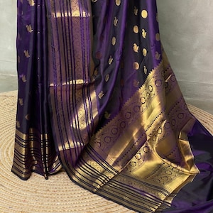 K&ouml;nnte beinhalten: Ein tiefvioletter Sari mit einer breiten, verzierten goldenen Bord&uuml;re mit komplexen geometrischen und floralen Mustern. Der Stoff ist mit kleinen, verstreuten goldenen Motiven verziert, die einen reichen Kontrast zur dunkelvioletten Basis bilden.
