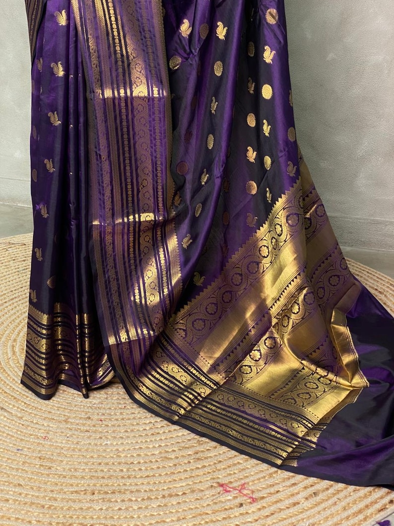 K&ouml;nnte beinhalten: Ein tiefvioletter Sari mit einer breiten, verzierten goldenen Bord&uuml;re mit komplexen geometrischen und floralen Mustern. Der Stoff ist mit kleinen, verstreuten goldenen Motiven verziert, die einen reichen Kontrast zur dunkelvioletten Basis bilden.
