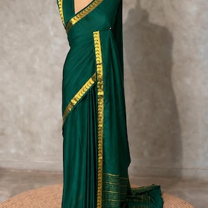 May include: A deep emerald green saree featuring a prominent gold border with a repeating geometric pattern. The fabric has a smooth, satin-like finish, and the end piece displays horizontal gold stripes. The garment is draped on a mannequin.