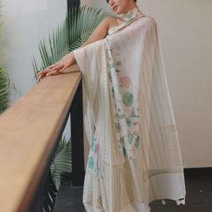 May include: An off-white saree featuring a delicate floral print with pink lotus flowers and green leaves. The fabric is adorned with vertical gold metallic stripes and a wide, textured gold border. The garment includes a matching high-neck collar.